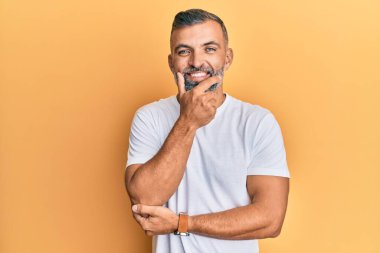 Orta yaşlı beyaz tişört giyen yakışıklı bir adam kameraya kendinden emin bakıyor kollarını kavuşturmuş ve çenesini kaldırmış gülümsüyor. Olumlu düşünmek. 