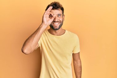 Sarı arka planda sıradan sarı tişörtler giyen sakallı yakışıklı bir adam. El hareketleriyle gülümsüyor, gözleriyle mutlu yüzle parmaklarının arasından bakıyor.. 