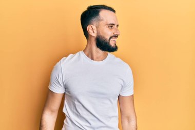 Sakallı, sıradan beyaz tişörtlü, yüzünde gülümseme ve doğal ifadeyle yan yana bakan genç bir adam. Kendinden emin gülen. 