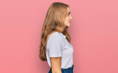 Beyaz günlük tişört giyen genç beyaz bir kız. Yan tarafa bakıyor, rahat bir poz veriyor. Kendine güvenen bir gülümsemesi var.. 