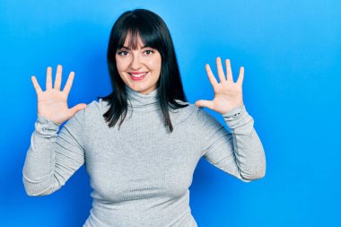 Genç İspanyol kadın gündelik giysiler giyer ve parmaklarını on numara ile gösterir ve gösterirken kendinden emin ve mutlu gülümser.. 