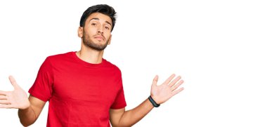 Gündelik kırmızı tişört giyen, elleri ve kolları havada, kafası karışmış genç ve yakışıklı bir adam. şüphe kavramı. 