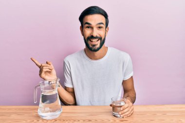 Genç İspanyol adam masaya oturmuş su içiyor. Gülümsüyor, mutlu bir şekilde elini ve parmağını yana doğru gösteriyor. 