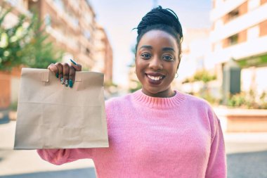 Genç Afro-Amerikalı kadın gülümseyerek elinde kağıt torbayla şehirde yiyecek taşıdığını söylüyor..