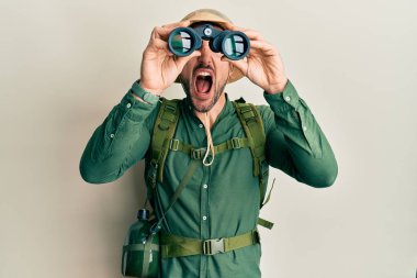 Handsome man with beard wearing explorer hat looking through binoculars angry and mad screaming frustrated and furious, shouting with anger looking up. 