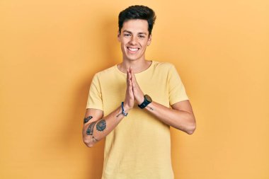 Gündelik sarı tişört giyen genç İspanyol adam elleriyle dua edip af diliyor kendinden emin bir şekilde gülümsüyor.. 