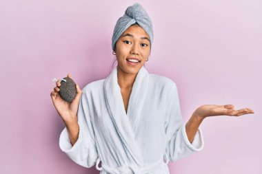 Duş havlusu şapkası ve bornoz giyen genç Çinli kadın başarıyı kutlamak için sünger taşı taşıyor mutlu bir gülümsemeyle ve kazanan yüz ifadesiyle elini kaldırarak. 