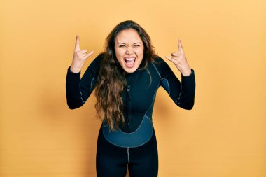 Young hispanic girl wearing diver neoprene uniform shouting with crazy expression doing rock symbol with hands up. music star. heavy music concept. 