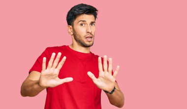 Kırmızı tişört giyen genç yakışıklı adam korku ifadesinden korkuyor ve dehşete düşüyor. El hareketlerini durdur, şok içinde bağır. panik konsepti. 