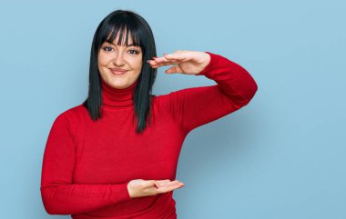 Latin kökenli genç bir kadın, gündelik kıyafetler giyiyor. Elleriyle işaret ediyor. Ölçü sembolü. Kameraya bakıp gülümsüyor. ölçüm kavramı. 