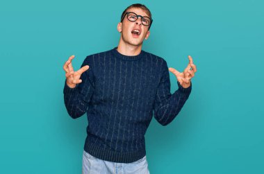 Young blond man wearing casual clothes and glasses crazy and mad shouting and yelling with aggressive expression and arms raised. frustration concept. 