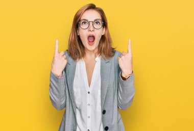 İş stili ve gözlük takan genç beyaz kadın şaşkın şaşkın yukarı bakıyor ve parmaklarıyla işaret ediyor ve kollarını kaldırıyor.. 
