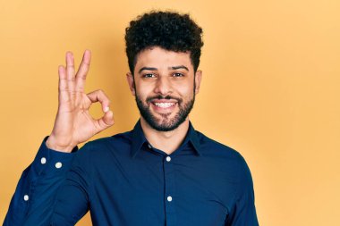 Young arab man with beard wearing casual shirt smiling positive doing ok sign with hand and fingers. successful expression. 