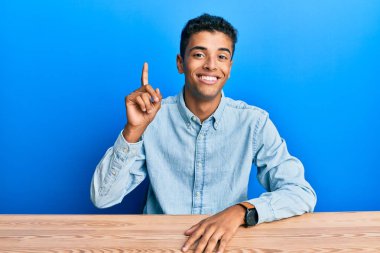 Genç, yakışıklı, Afro-Amerikalı, gündelik giysiler içinde masanın üzerinde oturuyor ve bir numaralı parmağını göstererek gülümsüyor.. 