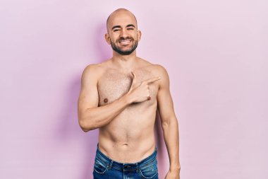 Genç kel adam, yüzünde mutlu ve doğal bir ifadeyle elleri ve parmaklarıyla gülümseyen bir gülümsemeyle üstsüz bir şekilde ayakta duruyor. 