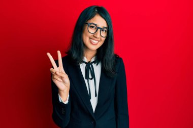 Güzel Asyalı genç bayan iş elbisesi giyiyor ve iki numaralı parmaklarıyla işaret ederken kendinden emin ve mutlu gülümsüyor.. 