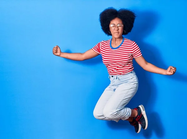 若い美しいアフリカ系アメリカ人女性のカジュアルな服や眼鏡を着て幸せな笑顔 孤立した青の背景の上に笑顔でジャンプ — ストック写真