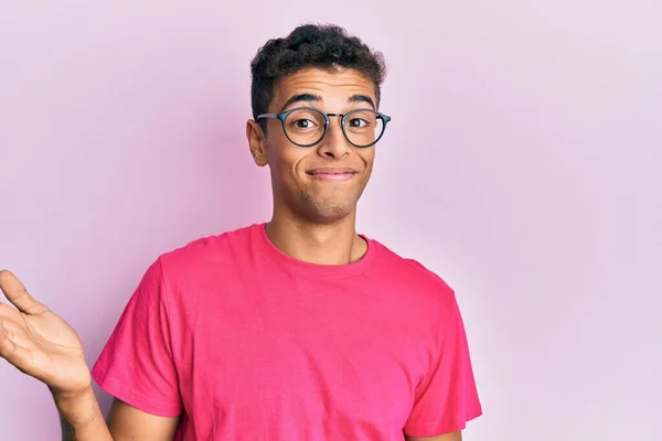 年轻英俊的非洲裔美国男子 戴着眼镜罩在粉色的背景上 毫无头绪 满脸困惑 手臂和双手高举 怀疑概念 — 图库照片