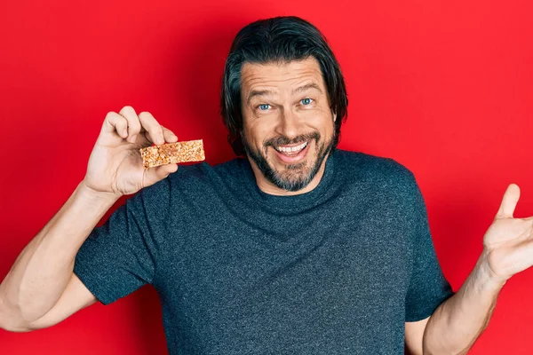 Middle age caucasian man eating protein bar as healthy energy snack celebrating achievement with happy smile and winner expression with raised hand