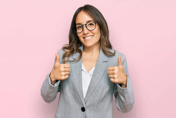 若いブルネットの女性は ビジネス服の成功サインを身に着けている手で積極的なジェスチャーを行う 親指を笑顔と幸せ 陽気な表情と勝者のジェスチャー — ストック写真