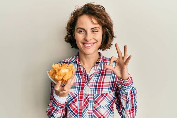 Młoda Brunetka Trzyma Chipsy Robi Znak Palcami Uśmiechając Przyjazny Gest — Zdjęcie stockowe