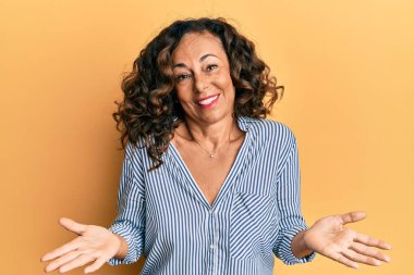 Orta yaşlı İspanyol kadın gündelik kıyafetler giyiyor, elleri ve kolları havada, kafası karışmış bir ifade takınıyor. şüphe kavramı. 