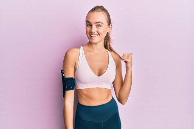Beautiful blonde woman wearing sportswear and arm band smiling with happy face looking and pointing to the side with thumb up. 