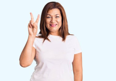 Orta yaşlı İspanyol bir kadın izole edilmiş arka planda durup kameraya bakıp zafer işareti yaparken gülümsüyor. sayı