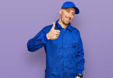 Bald man with beard wearing builder jumpsuit uniform doing happy thumbs up gesture with hand. approving expression looking at the camera showing success. 