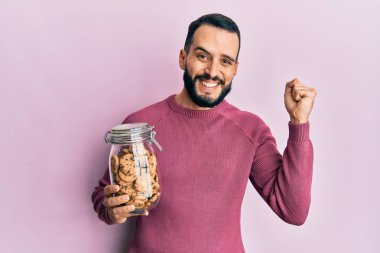 Sakallı genç adam elinde çikolatalı kurabiye kavanozuyla gururla bağırıyor, zaferi kutluyor ve başarıyı kollarını kaldırarak kutluyor. 