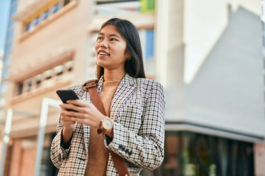 Genç Asyalı iş kadını şehirde akıllı telefon kullanarak mutlu bir şekilde gülümsüyor..