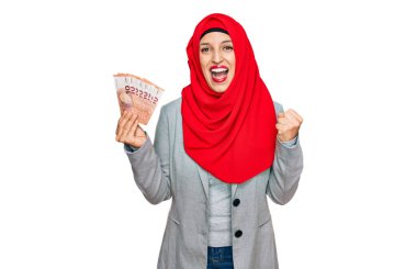 Beautiful hispanic woman wearing islamic hijab holding 10 euros banknotes screaming proud, celebrating victory and success very excited with raised arms 