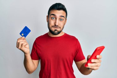 Young hispanic man holding smartphone and credit card puffing cheeks with funny face. mouth inflated with air, catching air. 