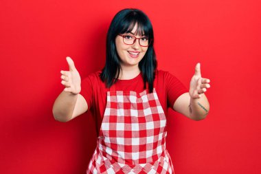 Genç İspanyol kadın aşçı önlüğü ve gözlük takıp kameraya bakıyor. Sarılmak için kollarını açmış gülümsüyor. Mutluluğu kucaklayan neşeli bir ifade.. 