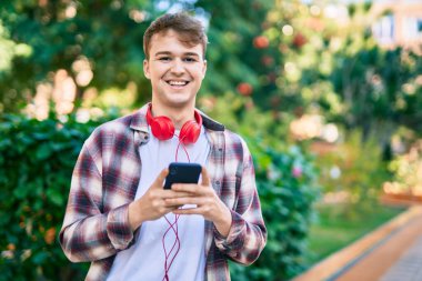 Parkta akıllı telefon ve kulaklık kullanarak gülümseyen beyaz bir genç..