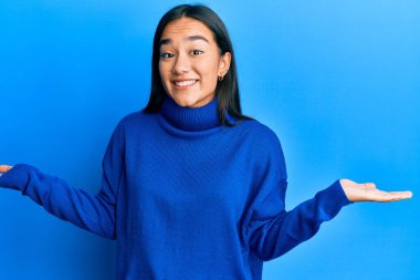 Genç Asyalı kadın, sıradan bir kazak giyip gülümsüyor. İki elini de açık tutuyor. Karşılaştırma ve reklam karşılaştırması yapıyor ve dengeyi sağlıyor. 