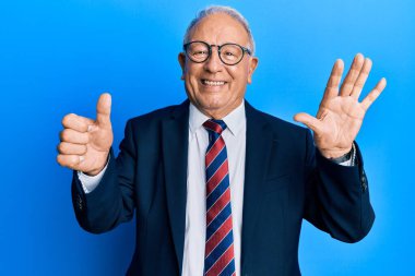 Beyaz üst düzey bir adam iş elbisesi ve kravat takıyor. Altıncı parmaklarını gösterip işaret ederken kendinden emin ve mutlu gülümsüyor.. 