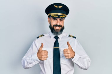 İspanyol asıllı genç adam uçak pilotu üniforması giyiyor. Başarı işareti el hareketleriyle olumlu bir hareket yapıyor. Gülümsüyor ve mutlu. Neşeli ifade ve kazanan jest. 