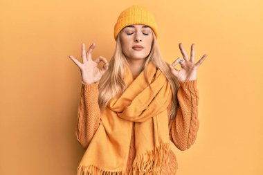 Kafkasyalı genç bir kadın yün kazak ve kep giyiyor rahatlıyor ve gözleri kapalı gülümsüyor parmaklarıyla meditasyon hareketi yapıyor. yoga konsepti. 