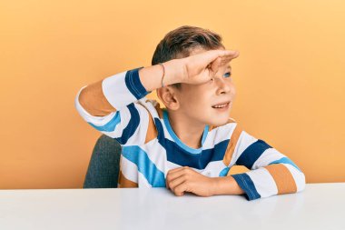 Kafkasyalı sevimli bir çocuk, gündelik giysiler giymiş masada mutlu bir şekilde oturuyor ve gülümsüyor. arama kavramı. 