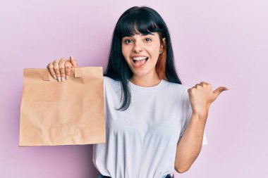 Elinde kağıt torba tutan genç İspanyol kız başparmağıyla yan tarafı işaret ediyor mutlu bir şekilde ağzı açık gülümsüyor. 