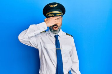 Sakallı, gri saçlı, uçak pilotu üniforması giyen, yüzünü ve gözlerini kapayarak utangaç ifadesiyle parmaklarının arasından bakan orta yaşlı bir adam.. 