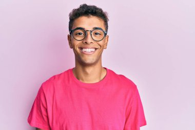Genç, yakışıklı, Afro-Amerikan bir adam pembe arka planda gözlük takıyor ve yüzünde mutlu ve havalı bir gülümseme var. şanslı kişi. 