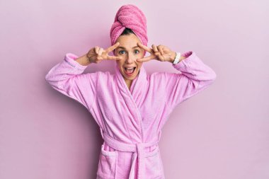 Duş başlığı ve bornoz takan genç sarışın kadın barış sembolü yapıyor. Yüzünde parmaklar, gülümseyerek zaferi gösteriyor. 