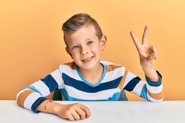 Beyaz elbiseli sevimli bir çocuk masanın üzerine oturmuş iki numarayı işaret ediyor ve kendinden emin ve mutlu bir şekilde gülümsüyor.. 