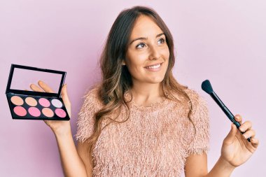 Makyaj fırçası ve allık tutan genç esmer bir kadın. Gülümsüyor, yan tarafa bakıyor ve düşünüyor.. 