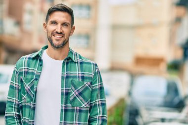 Genç beyaz adam gülümseyerek şehirde mutlu mesut duruyor..