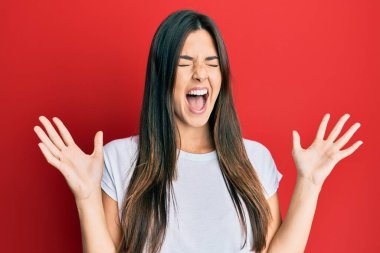 Genç esmer kadın kırmızı arka plan üzerine gündelik beyaz tişört giyerek çılgın ve başarıyı kollarını kaldırarak ve gözleri kapalı heyecanla bağırarak kutluyor. Kazanan konsept 