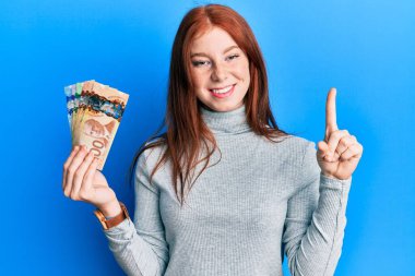 Kanada doları tutan genç kızıl saçlı kız bir fikir ya da soruyla gülümsüyor ve mutlu bir yüzle işaret ediyor. 