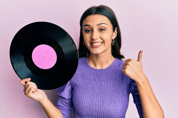 Young hispanic woman holding vinyl disc smiling happy and positive, thumb up doing excellent and approval sign